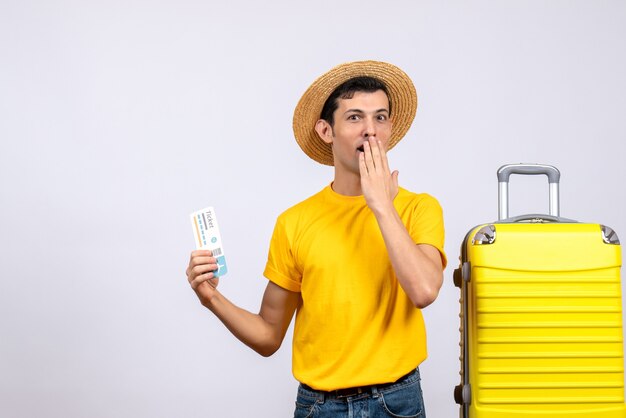 Vista frontal confusa jovem turista em pé perto de uma mala amarela segurando uma passagem.