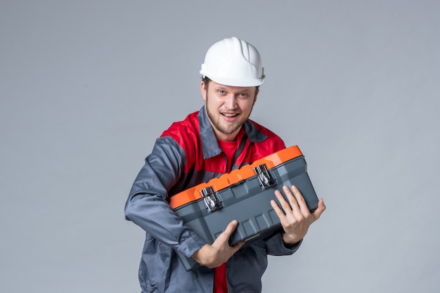 Vista frontal, construtor masculino uniformizado segurando uma caixa de ferramentas pesada em fundo cinza
