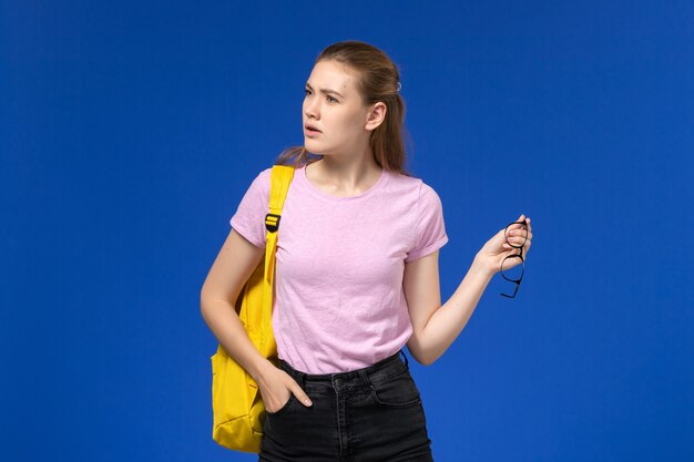 Vista frontal da aluna com camiseta rosa e mochila amarela posando na parede azul