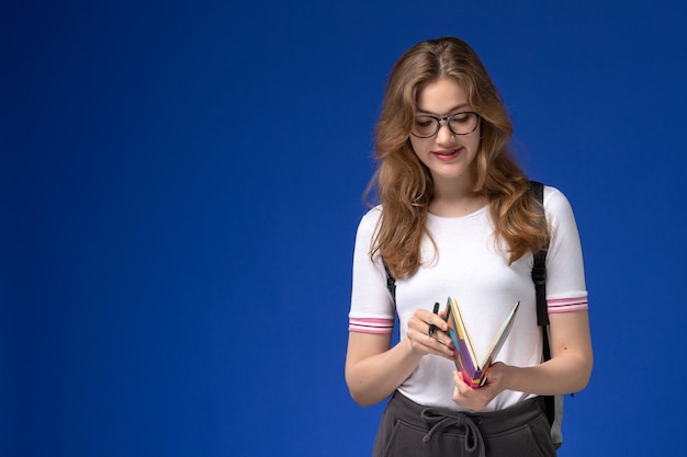 Vista frontal da aluna de camisa branca segurando uma caneta e um caderno na parede azul