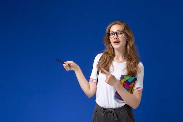Vista frontal da aluna de camisa branca segurando uma caneta e um caderno na parede azul