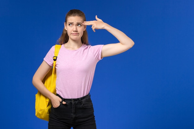 Vista frontal da aluna em uma camiseta rosa com mochila amarela posando na parede azul claro