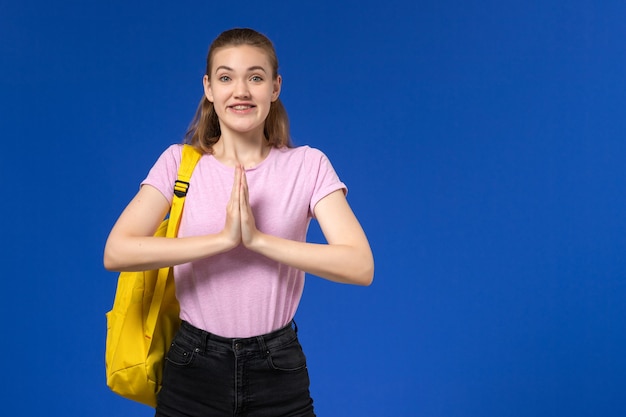 Vista frontal da aluna em uma camiseta rosa com mochila amarela sorrindo na parede azul clara