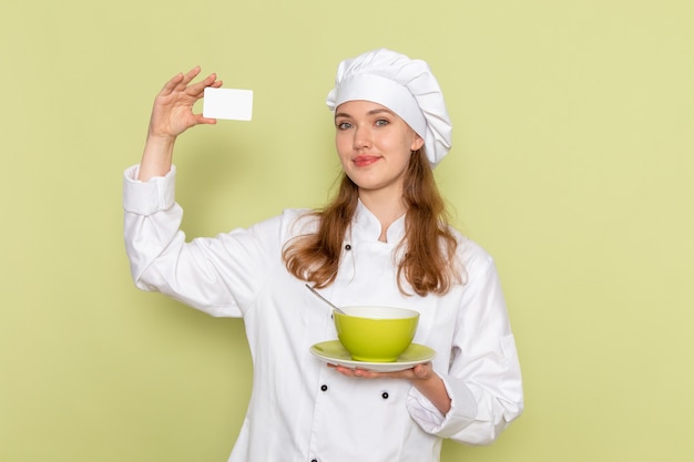 Vista frontal da cozinheira de terno branco segurando o cartão e o prato na parede verde