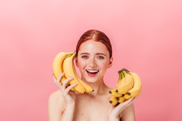 Vista frontal da deslumbrante garota gengibre com bananas. foto de estúdio de feliz mulher nua segurando frutas tropicais em fundo rosa.