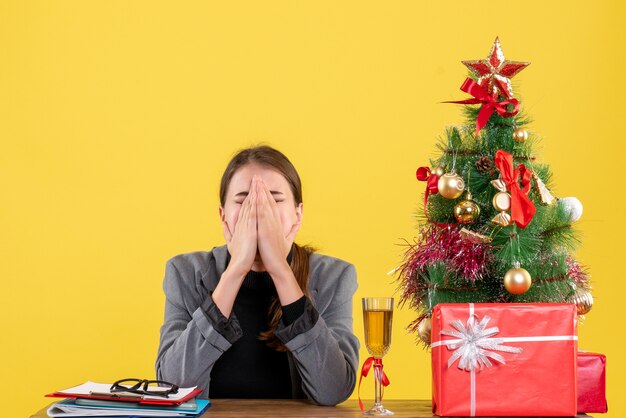 Vista frontal da garota triste sentada na mesa gritando e cobrindo o rosto com as mãos da árvore de natal e coquetel de presentes