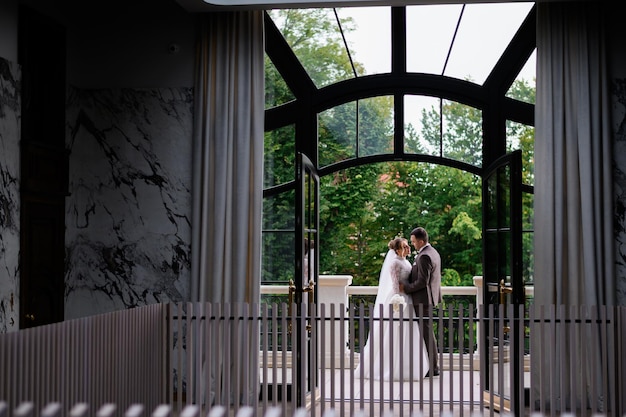 Vista frontal da janela grande e transparente com porta aberta fora da qual recém-casados posando e abraçando na varanda durante o dia do casamento