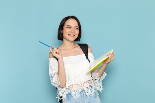 Vista frontal da jovem aluna de camisa branca jeans e bolsa preta escrevendo notas sobre o espaço azul.