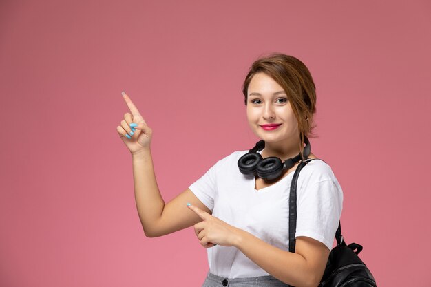 Vista frontal da jovem aluna em uma camiseta branca com bolsa e fones de ouvido posando e sorrindo sobre a mesa rosa.