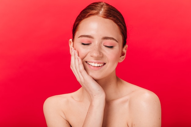 Vista frontal da jovem feliz, sorrindo com os olhos fechados. Menina ruiva nua posando sobre fundo vermelho.