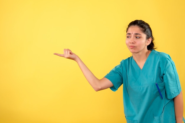 Vista frontal da jovem médica de uniforme apontando para a esquerda em pé na parede amarela