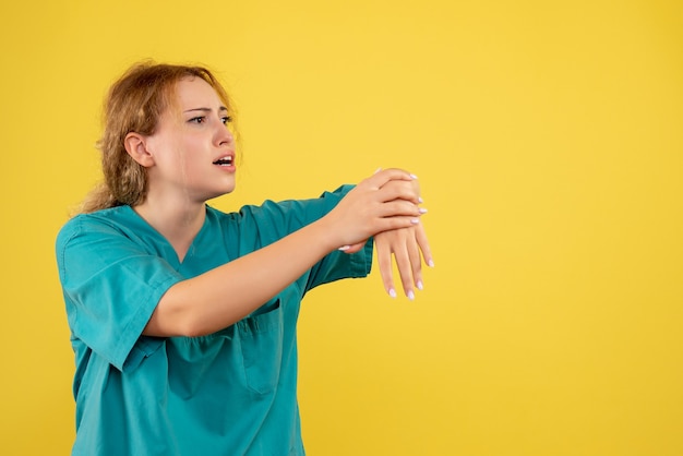 Vista frontal da jovem médica em camisa médica com o braço ferido na parede amarela