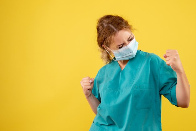 Vista frontal da jovem médica em traje médico e máscara estéril regozijando-se na parede amarela