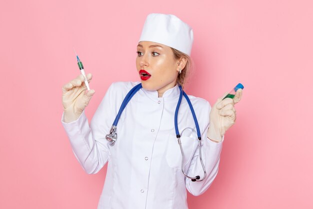 Vista frontal da jovem médica em um terno médico branco com um estetoscópio azul segurando a injeção e o frasco na saúde do hospital médico do espaço da luz.