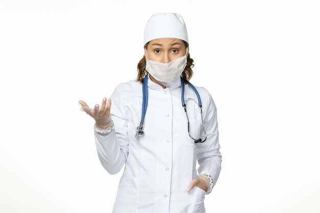 Vista frontal da jovem médica em um terno médico branco e com máscara devido a coronavírus na superfície branca