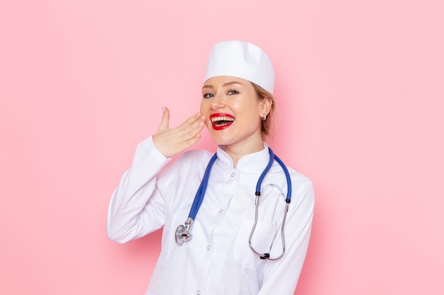 Vista frontal da jovem médica vestida de branco com estetoscópio azul posando e sorrindo no trabalho do espaço rosa