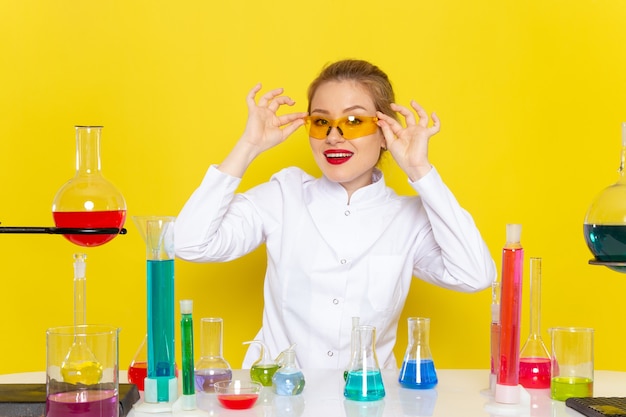 Vista frontal da jovem química feminina em um terno branco em frente à mesa com soluções de ed, sentada e sorrindo na química do espaço amarelo