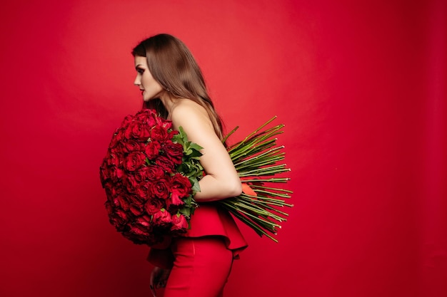 Vista frontal da linda mulher de vestido vermelho com maquiagem mantendo lindo buquê de rosas olhando para a câmera e sorrindo bonita modelo posando em fundo vermelho isolado conceito de dia dos namorados