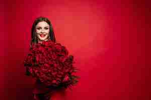 Foto grátis vista frontal da linda mulher de vestido vermelho com maquiagem mantendo lindo buquê de rosas olhando para a câmera e sorrindo bonita modelo posando em fundo vermelho isolado conceito de dia dos namorados