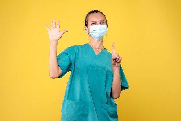 Vista frontal da médica em traje médico e máscara estéril na parede amarela