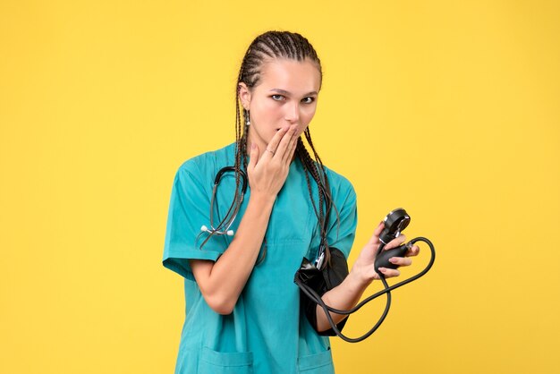 Vista frontal da médica em traje médico, verificando sua pressão na parede amarela