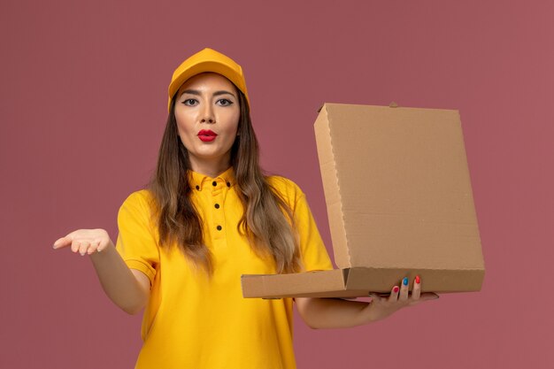 Vista frontal da mensageira de uniforme amarelo e boné segurando a caixa de comida aberta na parede rosa claro