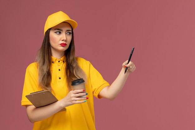 Vista frontal da mensageira de uniforme amarelo e boné segurando a xícara de café de entrega e o bloco de notas com caneta na parede rosa