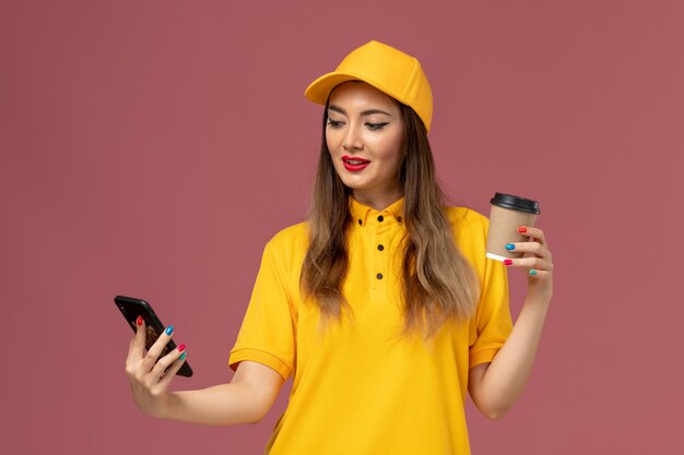 Vista frontal da mensageira de uniforme amarelo e boné segurando a xícara de café de entrega e o telefone na parede rosa