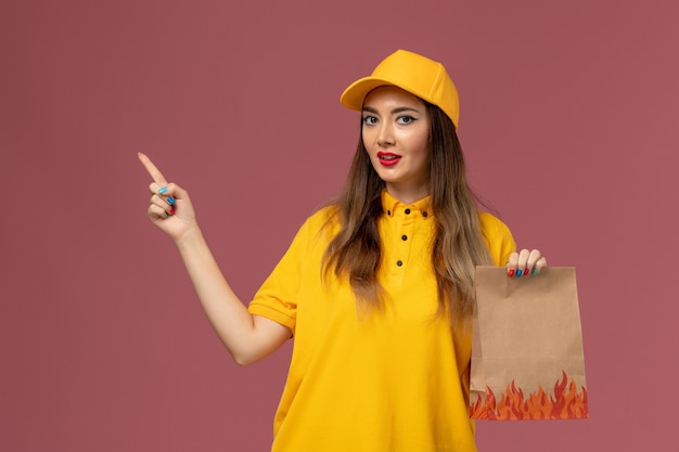 Vista frontal da mensageira de uniforme amarelo e boné segurando o pacote de alimentos na parede rosa claro