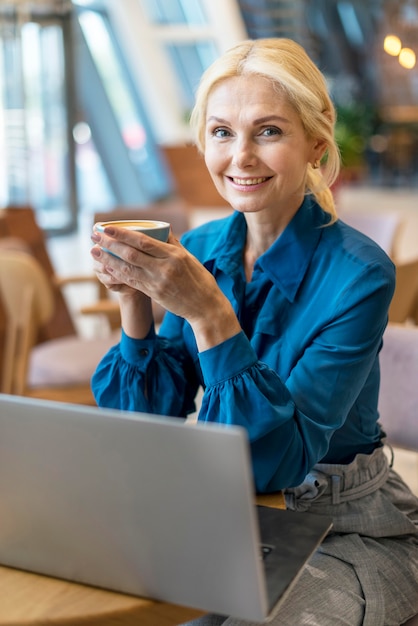 Vista frontal da mulher de negócios mais velha, tomando uma xícara de café e trabalhando no laptop