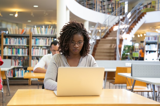 Vista frontal da mulher pensativa, trabalhando com o laptop na biblioteca