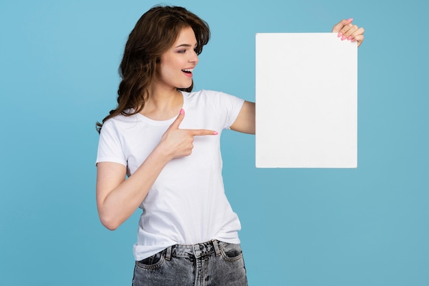 Vista frontal da mulher sorridente segurando e apontando para um cartaz em branco