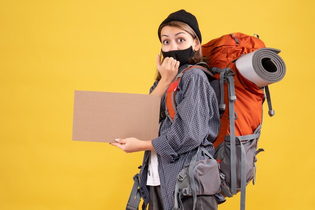 Vista frontal da mulher viajante com máscara preta e mochila segurando um papelão na parede amarela