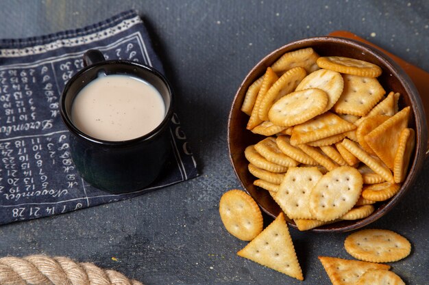 Vista frontal da placa marrom com biscoitos e batatas fritas, juntamente com o copo de leite na superfície cinza