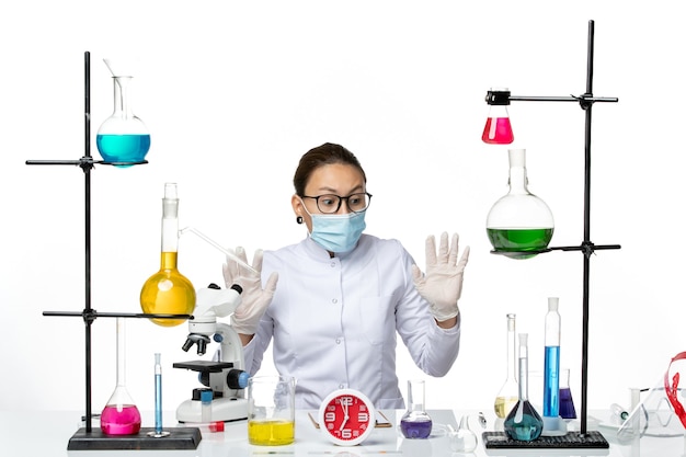 Vista frontal da química feminina em um terno médico branco com máscara sentada com soluções no chão branco.
