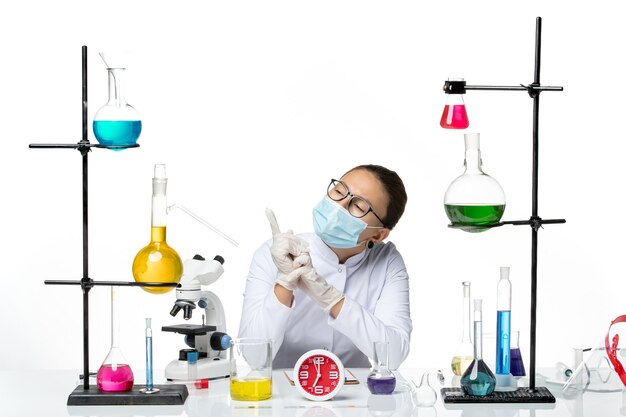 Vista frontal da química feminina em um terno médico branco com máscara sentada com soluções no laboratório de vírus covid-splash de químico de mesa branca