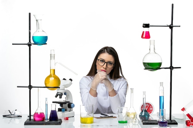 Vista frontal da química feminina em um terno médico branco sentada com diferentes soluções no laboratório de vírus pandêmico de ciência de mesa branca