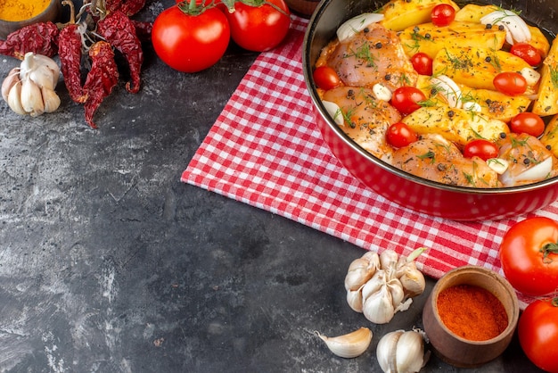 Vista frontal da refeição de frango cru com legumes de batatas em panela na toalha vermelha despojada e alhos de tomate no fundo cinza