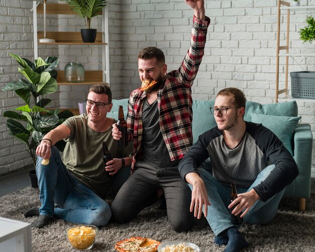 Vista frontal de alegres amigos do sexo masculino assistindo esportes na tv e comendo pizza