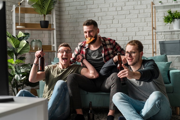 Vista frontal de alegres amigos do sexo masculino comendo pizza, cerveja e assistindo esportes na tv
