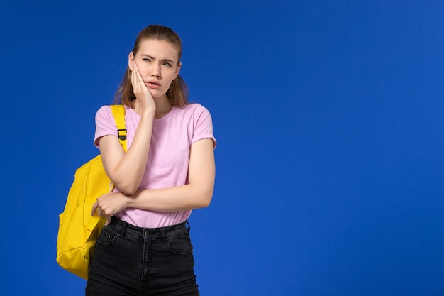 Vista frontal de aluna com camiseta rosa e mochila amarela pensando na parede azul-clara