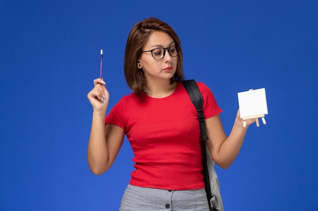 Vista frontal de aluna de camisa vermelha com mochila pintando um cavalete na parede azul