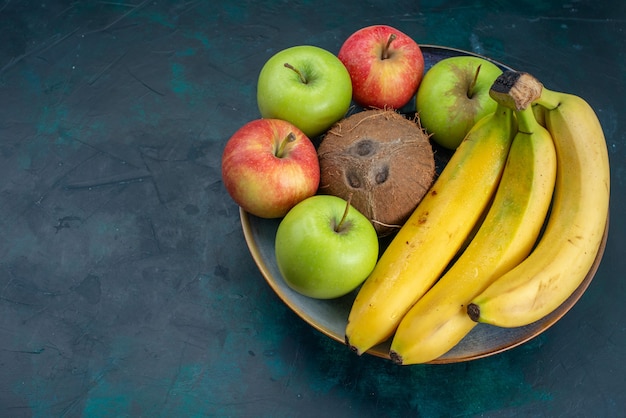 Vista frontal de diferentes composições de frutas, coco, maçãs e bananas na mesa azul-escuro frutas frescas exóticas suaves