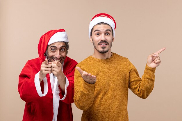 Vista frontal de dois amigos com chapéu de Papai Noel apontam o dedo mostrando a câmera em um fundo bege isolado