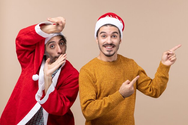 Vista frontal de dois amigos com chapéu de Papai Noel, um mostrando a direção com uma cara feliz em um fundo bege isolado
