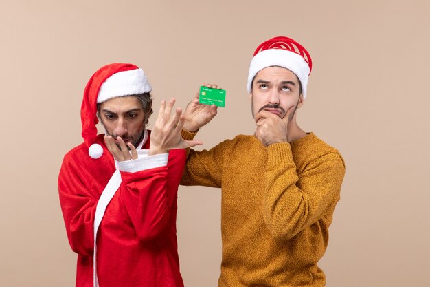 Vista frontal de dois amigos, um com um casaco de Papai Noel olhando para a mão dele e o outro segurando um cartão em um fundo bege isolado