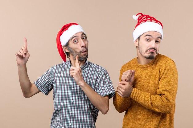 Vista frontal de dois caras confusos com chapéu de Papai Noel com um dedo apontando mostrando algo no fundo bege