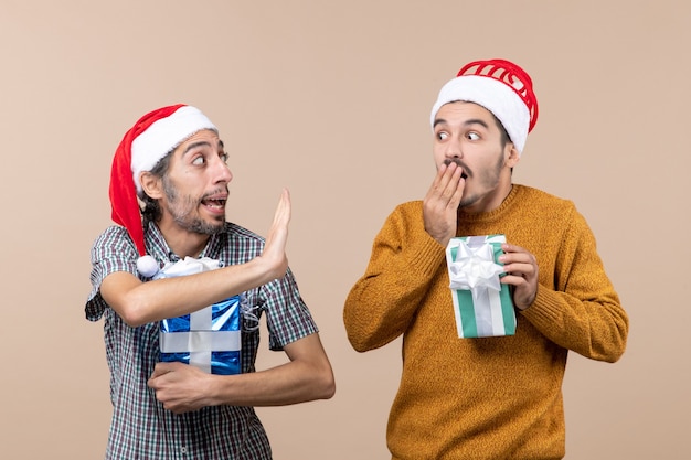 Vista frontal de dois caras confusos compartilhando segredo e segurando presentes de Natal em um fundo bege isolado