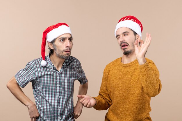 Vista frontal de dois caras de natal com chapéu de papai noel, um fazendo sinal de ok em fundo bege