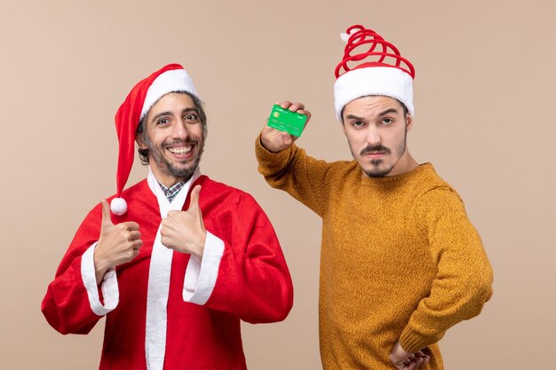 Vista frontal de dois caras felizes, um com casaco de papai noel e o outro com crédito colocando a mão na cintura em um fundo bege isolado